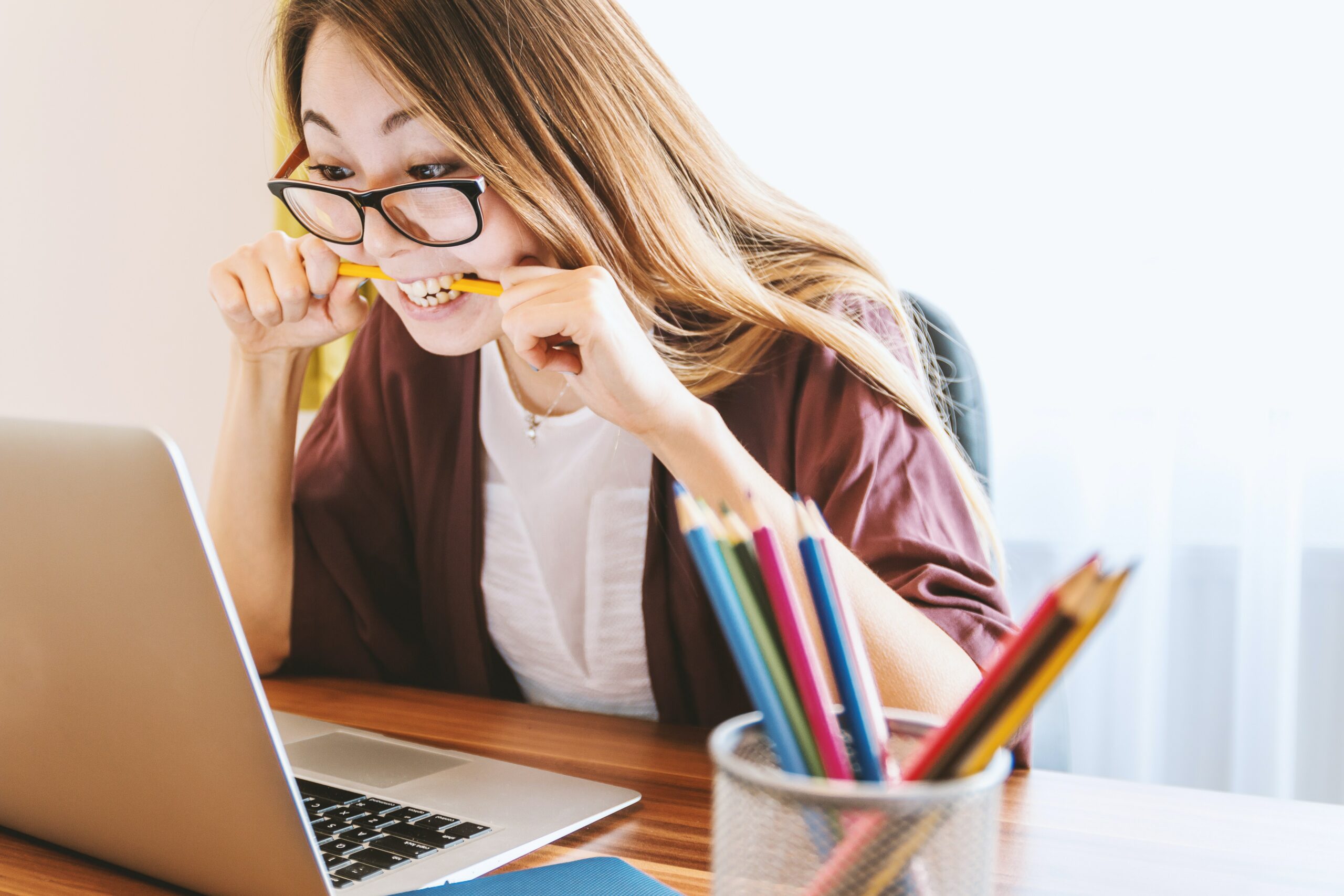 Young woman bites into pencil while eagerly staring at a laptop like other college graduates applying for jobs..